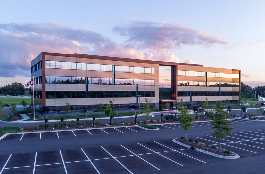 Modern three-story glass office building reflecting sunset hues; spacious, well-marked parking lot in foreground. Ideal for commercial renovation and construction projects, highlighting potential business growth opportunities.