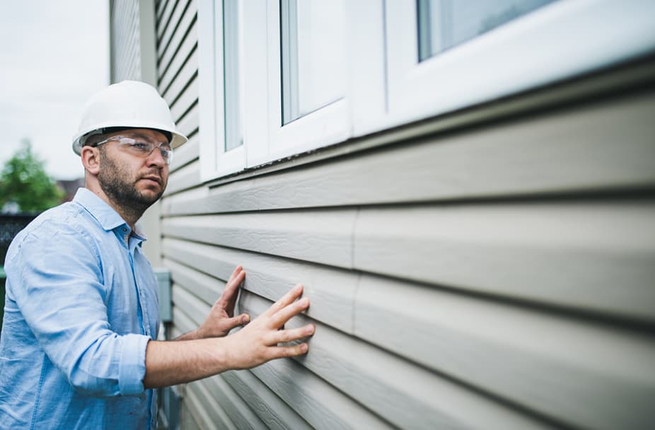 Construction expert in hard hat and glasses examining building siding for commercial renovation improvements.
