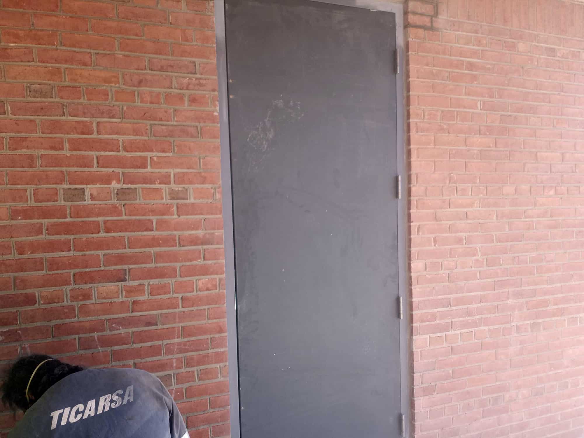 Metal door in brick wall with person in "TICARSA" shirt nearby, exterior renovation scene.