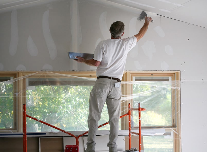 Person in white clothing skillfully repairs drywall with a trowel on ceiling; scaffolding setup; large windows covered in protective plastic sheeting.