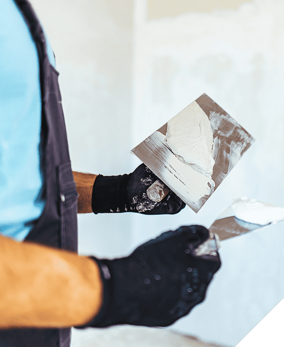Close-up of a person in black gloves holding two plastering trowels covered with white plaster, standing in a room with unfinished drywall, representing interior renovation services.