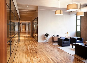 Modern office hallway with glass-walled rooms on one side and wooden flooring; features a seating area with two black chairs and a small desk. Stylish interior door installation for elegant workspace division, enhanced by hanging lights.