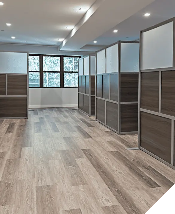 Empty office space with wooden commercial flooring, partitioned cubicles, and large sunlit windows.