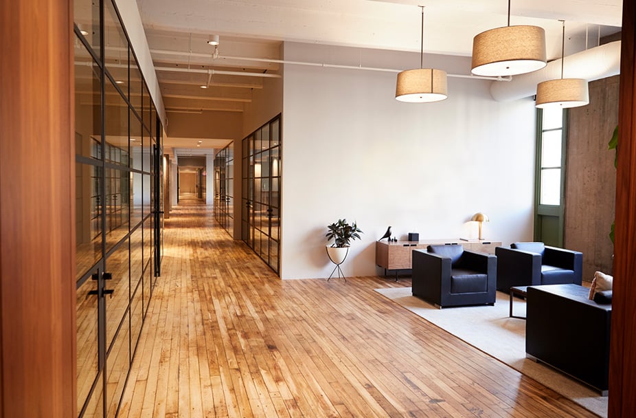 Office renovation with wooden floors and glass-walled meeting rooms. Sleek black armchairs in seating area, complemented by a modern small table and ceiling lights for optimal illumination.