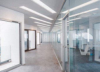 Image of a modern office hallway featuring glass walls and bright overhead lighting, with gray flooring leading to multiple rooms; ideal for showcasing professional interior renovation services and local handyman repairs for seamless efficiency.