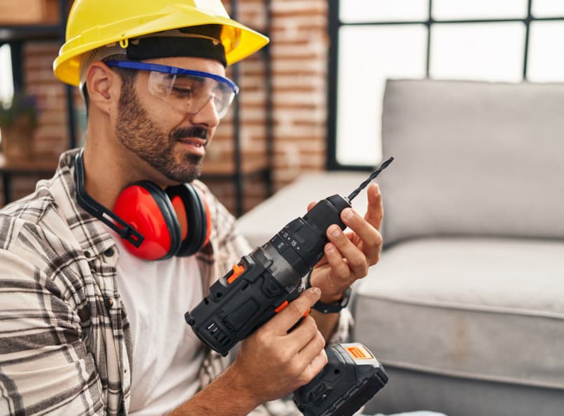 Handyman in safety gear adjusting power drill indoors, demonstrating expertise in interior and exterior renovation services.