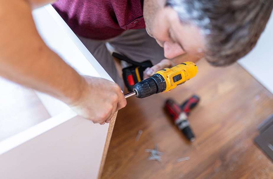 Person assembling white furniture using yellow electric drill on wooden floor during interior renovation, with screws and red drill nearby.
