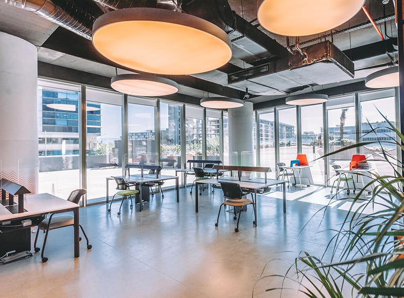 Modern office space with large windows, individual desks, black chairs, commercial lighting; colorful chair variations add vibrancy; visible outdoor buildings enhance ambience.