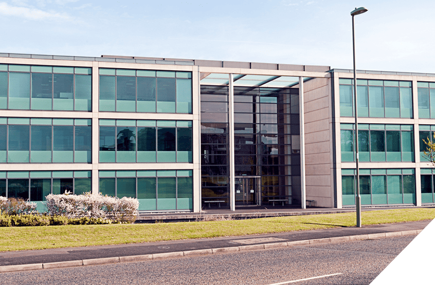 Modern three-story office building with green-tinted windows, central glass entrance, streetlamp, and grass verge. Exterior renovation and construction services.