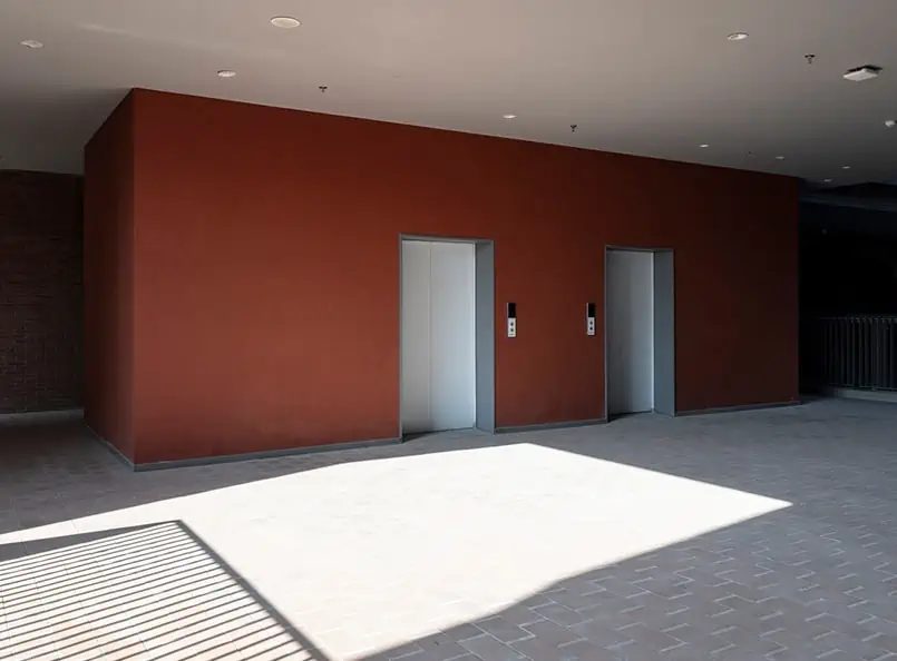 Elevator doors in a building with red wall and tile flooring, illuminated by sunlight, ideal for interior painting showcase.