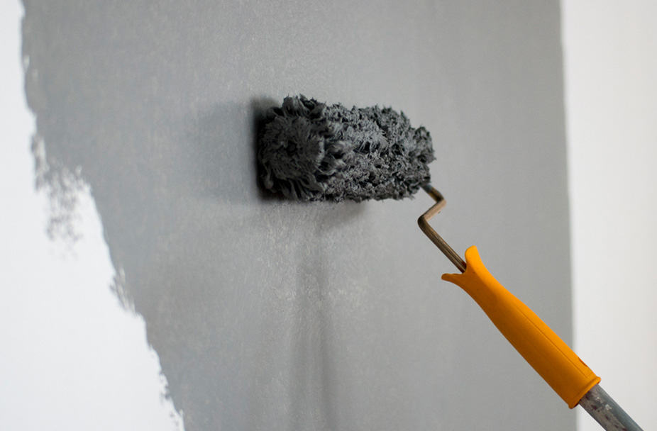 Yellow-handled paint roller applying gray paint to white wall for interior renovation project.