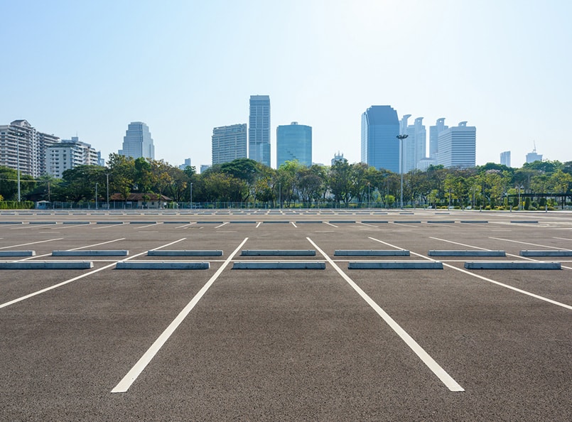 Empty parking lot with freshly painted white lines, surrounded by city skyscrapers and lush trees under a clear sky, ideal for efficient parking lot renovation services.