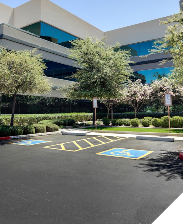 Modern office building with reflective windows; well-maintained accessible parking spaces; surrounding trees and shrubs under clear blue sky; optimized parking lot organization and maintenance.