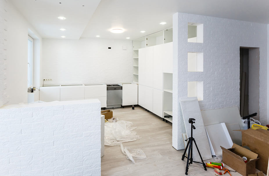 Interior kitchen remodel with white cabinets, unassembled furniture, and tools scattered on the floor; white walls and ceiling with recessed lighting.