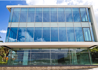 Three-story modern glass building with large windows, showcasing expert roofing craftsmanship under clear blue sky. Exterior renovation excellence.