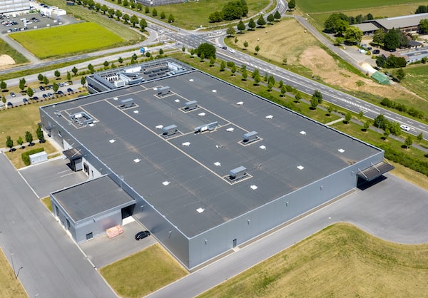 Aerial view of an expansive industrial building with a flat roof, surrounded by roads and green spaces, emphasizing the need for expert roofing services in large-scale renovations.