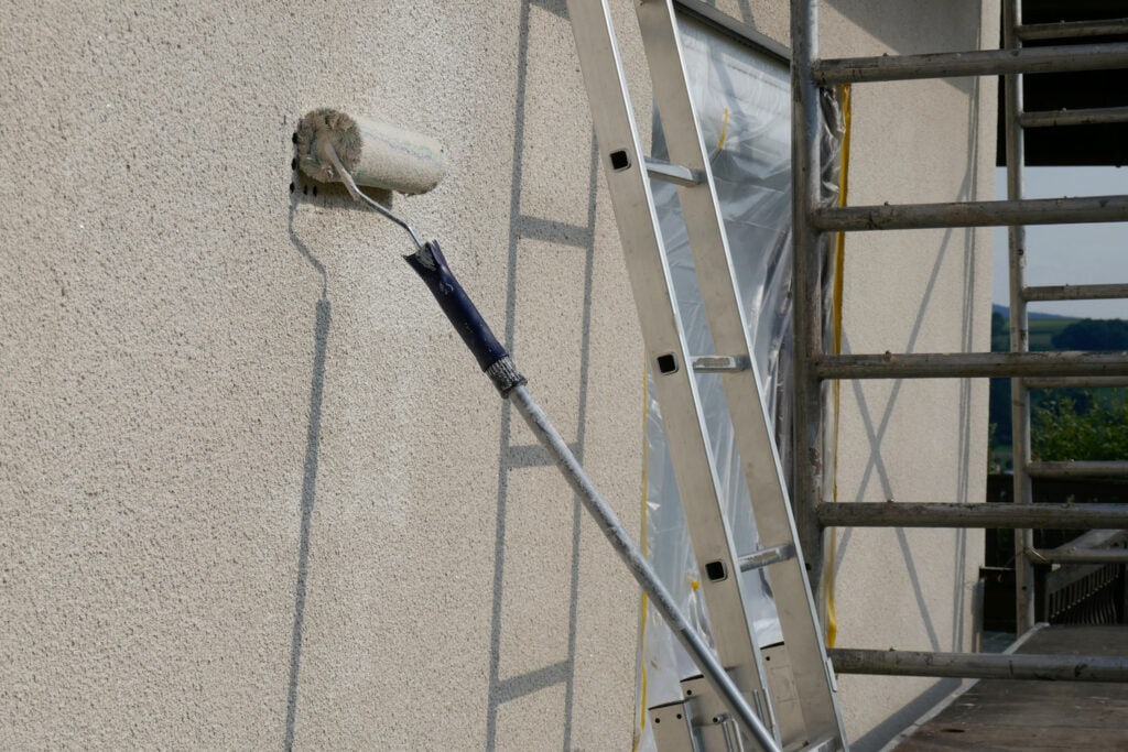Exterior painting using a paint roller on extension pole; ladder and scaffolding in background for building renovations.