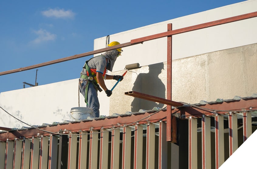 Worker in safety gear using roller brush for exterior wall painting on a sunny day.