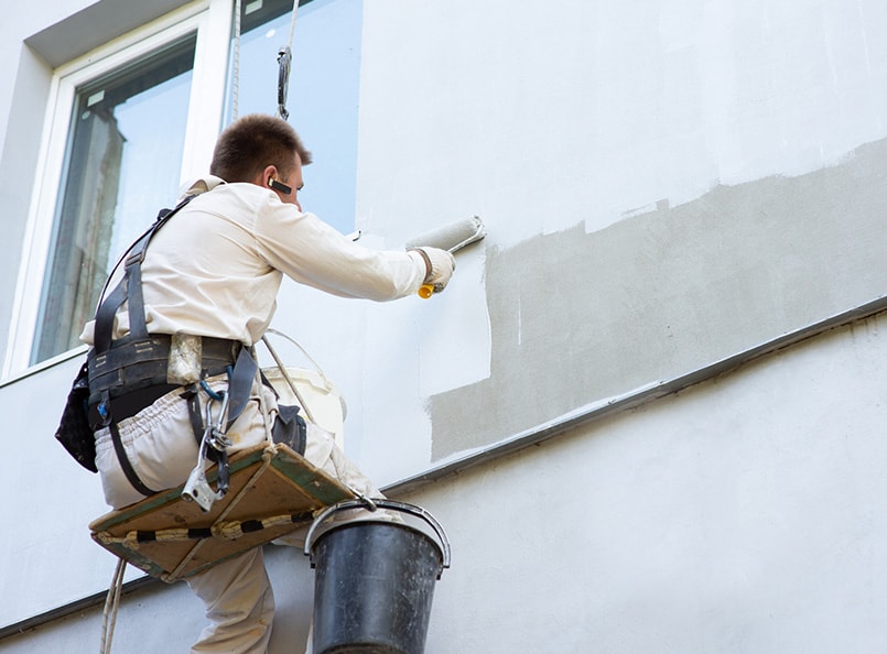 A construction worker in safety gear applies fresh exterior paint to a building using a roller on a suspended platform, with a paint bucket hanging beside.