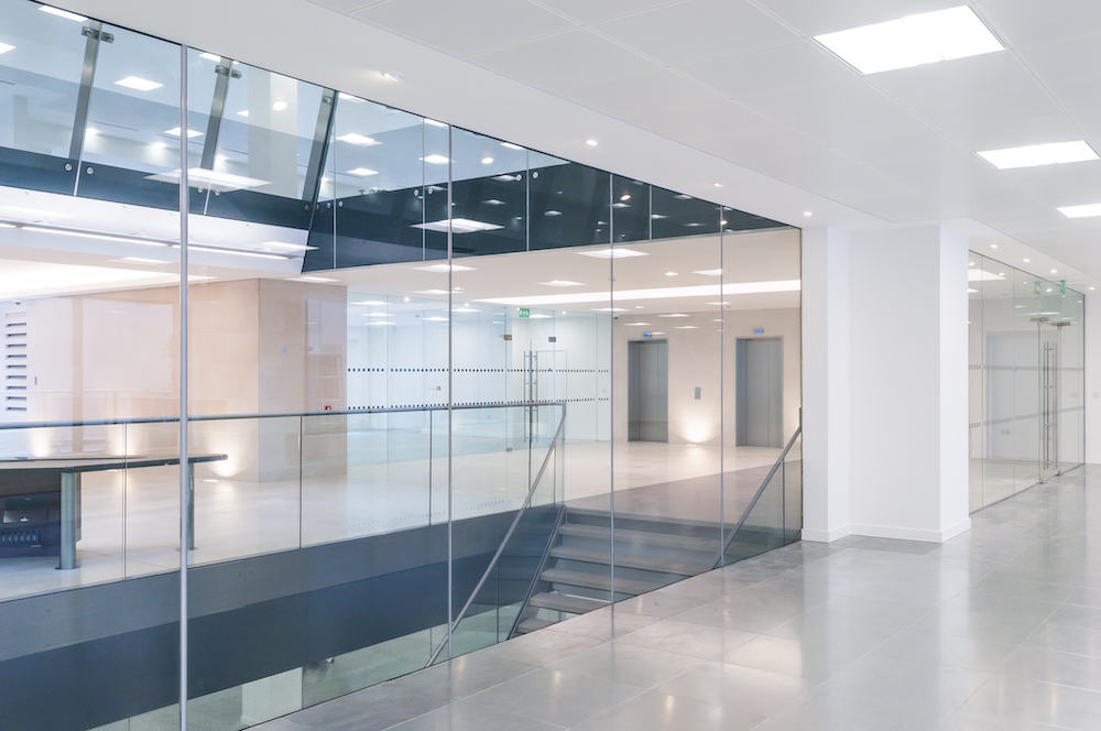 Modern office interior with glass walls, ceiling lights, empty hallway; includes two elevators and stairway to lower level; sleek design ideal for commercial glass repair projects.