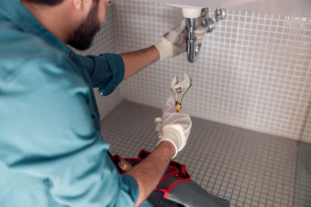 Gloved handyman using wrench for expert plumbing repair under bathroom sink with tiled backdrop.