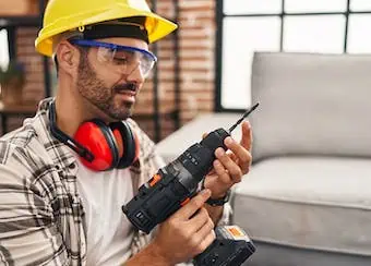 Construction worker in yellow hard hat using power drill confidently; wearing protective glasses and earmuffs; interior renovation setting.