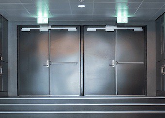 Grey double doors with push bars and illuminated exit signs, located at the top of a short staircase in a dim hallway; showcases recent interior renovations and remodels.