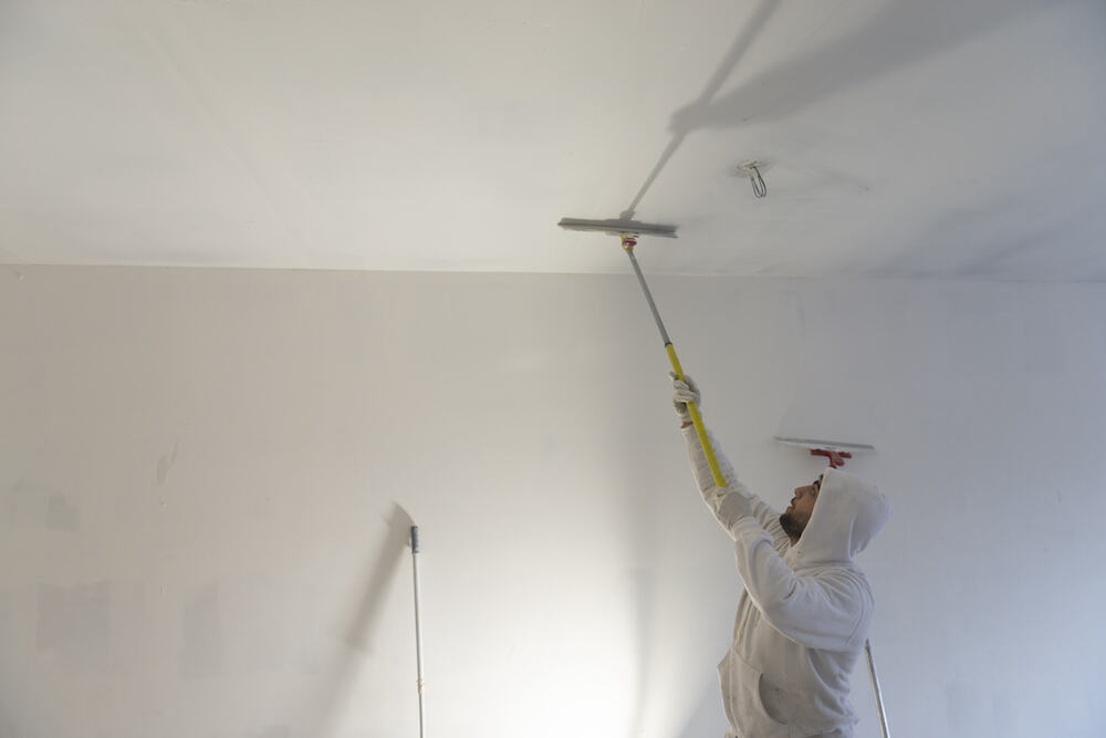 Expert craftsman in white hoodie skillfully painting white ceiling with long-handled tool, showcasing interior renovation services.