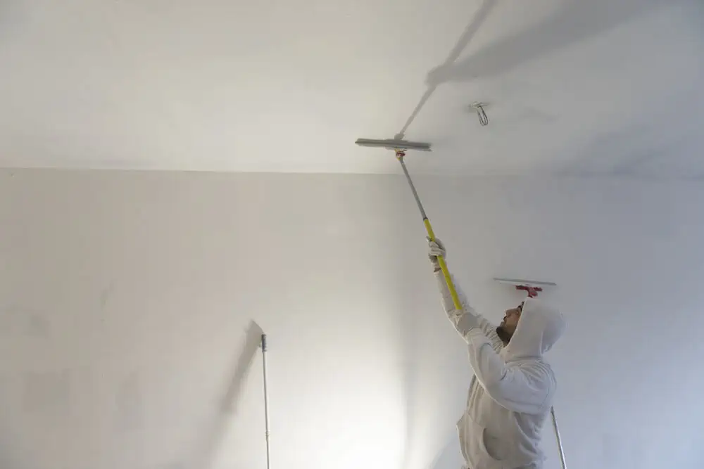 Expert craftsman in white hoodie skillfully painting white ceiling with long-handled tool, showcasing interior renovation services.