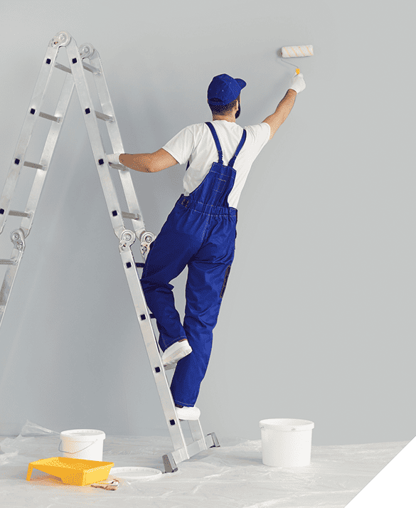 Interior painting process with a worker in blue overalls on a ladder, applying gray paint to the wall using a roller; neatly arranged paint supplies on the floor.