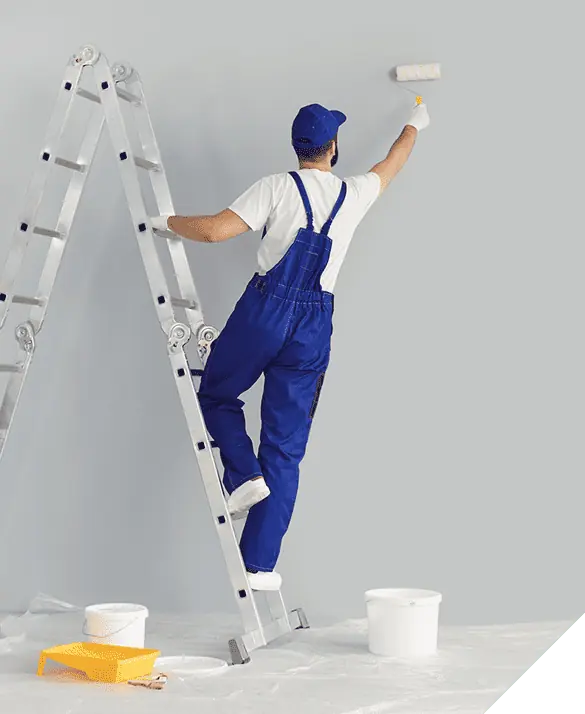 Interior painting process with a worker in blue overalls on a ladder, applying gray paint to the wall using a roller; neatly arranged paint supplies on the floor.