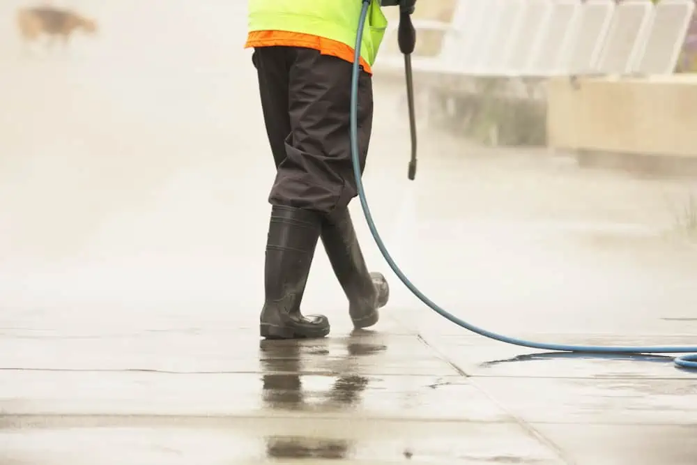 Construction worker using power washer to clean concrete surface, wearing high-visibility jacket and black boots, exterior renovation service.