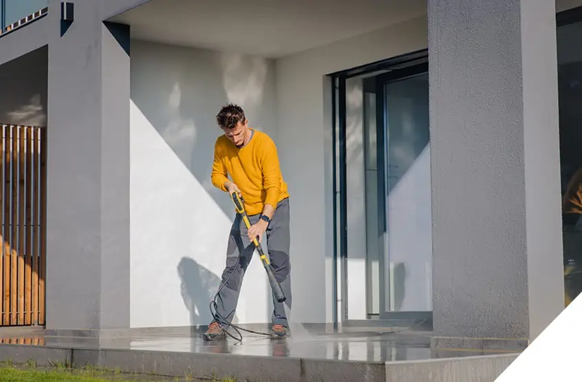 Image showing a person in a yellow sweater power washing a concrete patio outside a modern building, highlighting professional exterior cleaning renovation services.