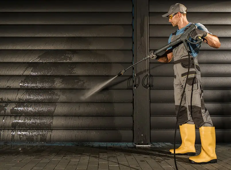 Worker power washing dark exterior wall, wearing safety gear and yellow boots for renovation.