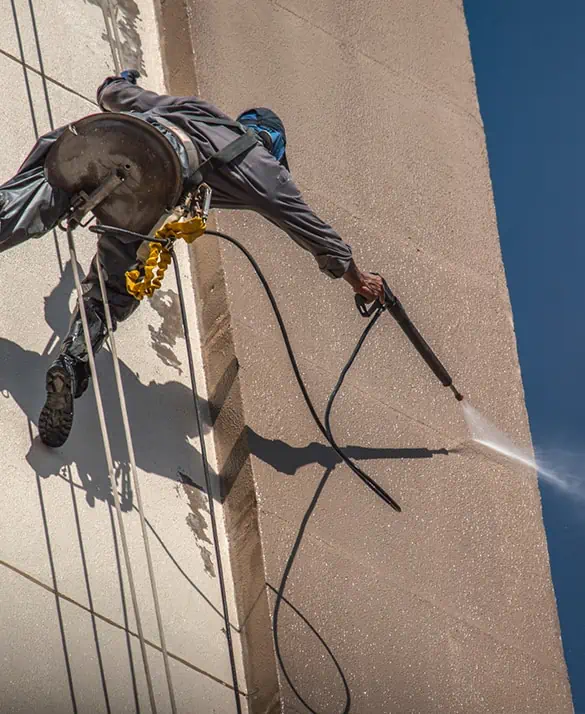 Worker using ropes to power wash high-rise building exterior, emphasizing professional renovation and cleaning services.