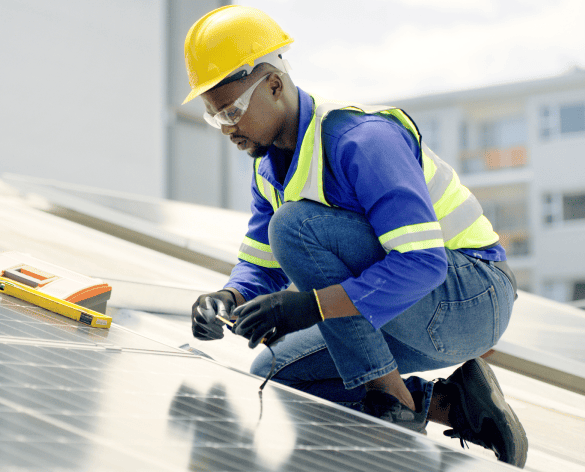 Worker in safety gear inspecting rooftop solar panel, wearing yellow helmet, gloves, and high-visibility vest; showcases professional roofing services with urban buildings in the background.