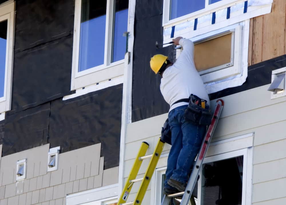 Construction worker in yellow hard hat installing siding and window on a building, showcasing professional renovation services.