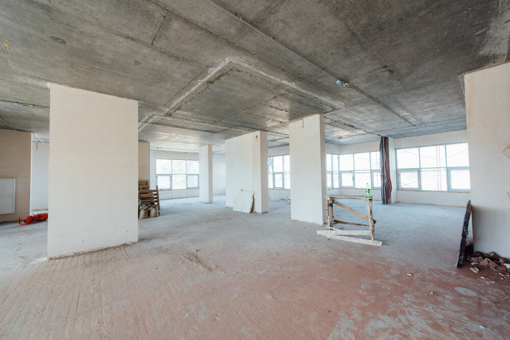 An unfinished room with concrete floors, concrete ceiling, large windows, scattered construction materials; ideal for expert roofing and renovation services.