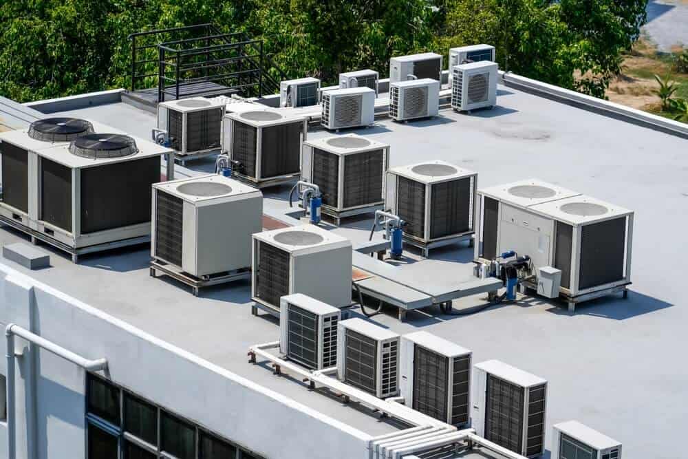 Aerial view of flat rooftop with multiple neatly arranged air conditioning units surrounded by greenery and pipes during a detailed commercial roof inspection.