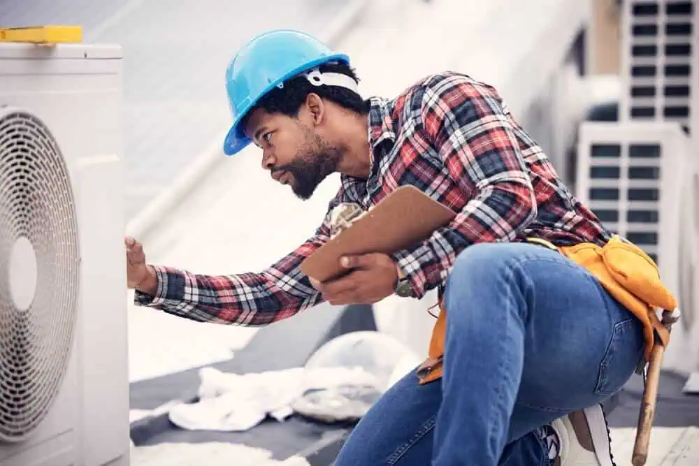 Construction worker in blue helmet and plaid shirt inspecting rooftop air conditioner unit with clipboard during commercial building envelope maintenance.