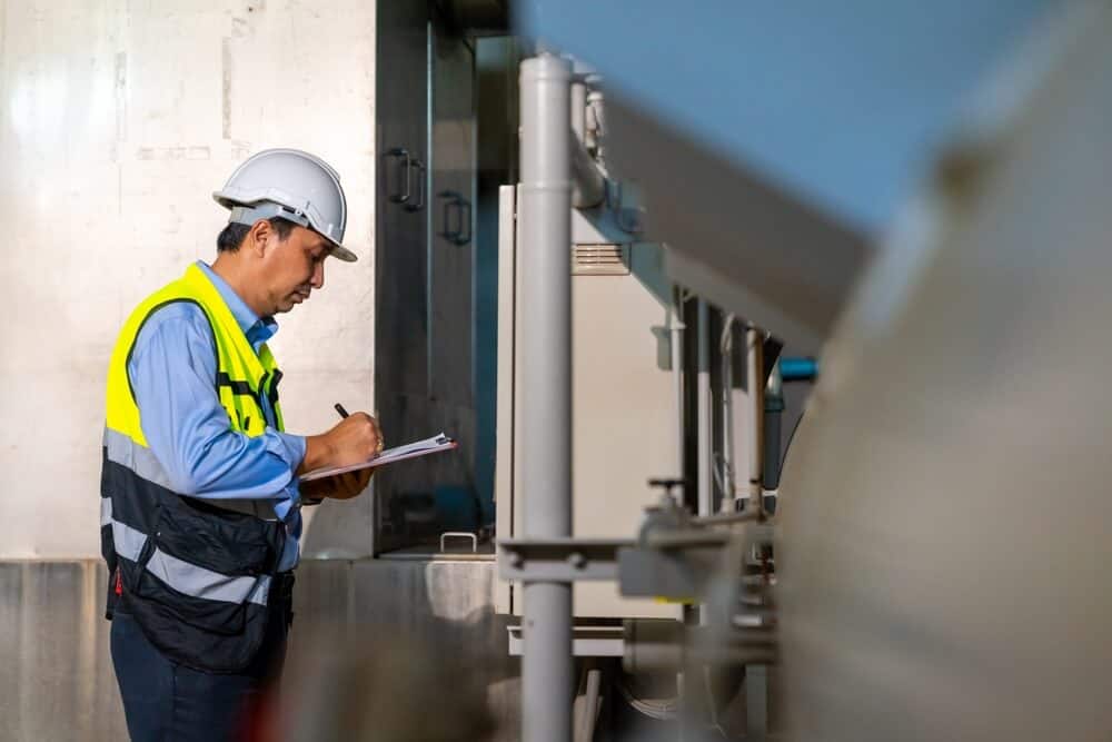 A construction worker in a hard hat and safety vest inspects industrial equipment, documenting details on a clipboard for commercial building envelope maintenance.