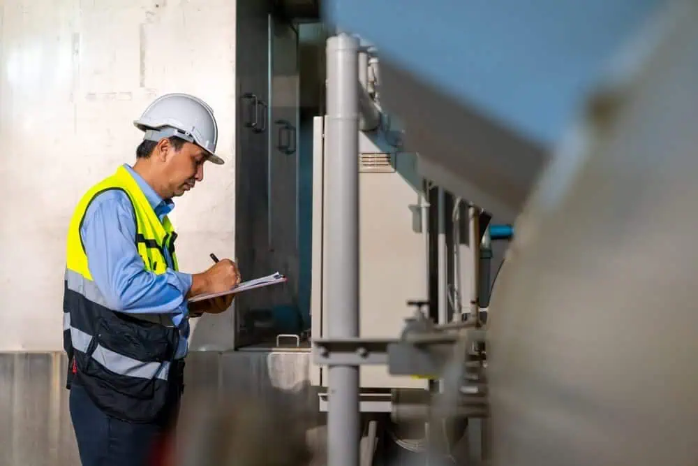 A construction worker in a hard hat and safety vest inspects industrial equipment, documenting details on a clipboard for commercial building envelope maintenance.