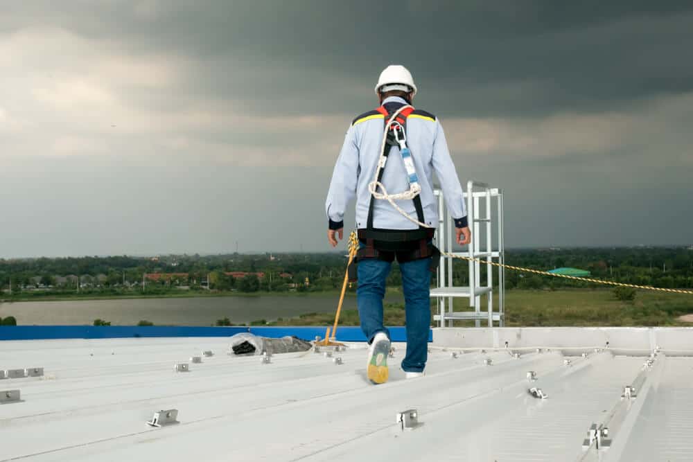 Worker in safety gear on rooftop, wearing harness for commercial roof inspection against overcast sky and distant landscape backdrop.