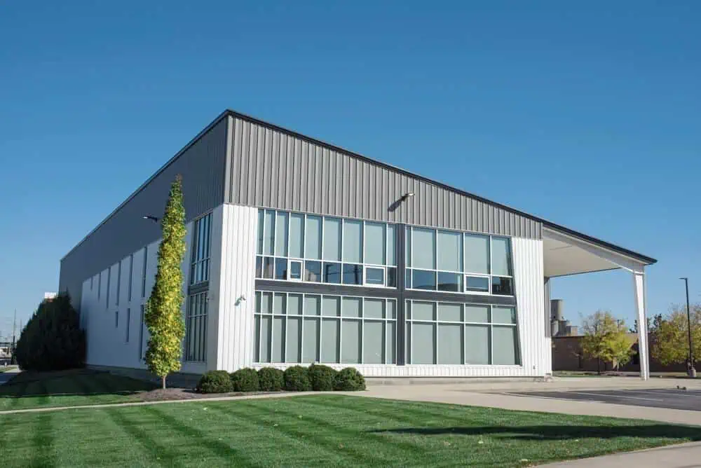 Contemporary industrial building with large windows, sleek commercial siding, flat roof; manicured lawn and tall narrow tree in landscaped exterior against clear blue sky.