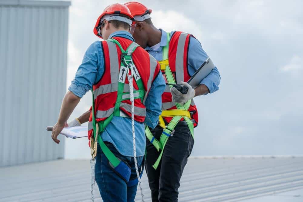 Two construction workers in safety gear inspect commercial roof, analyzing blueprints and digital plans for renovation services.