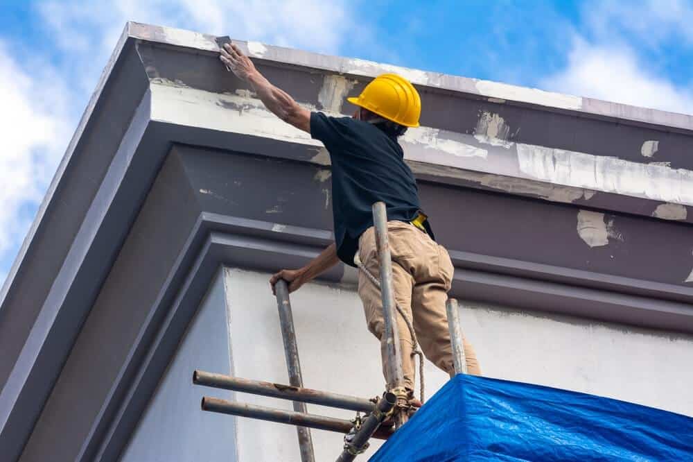 Construction worker in yellow hard hat on scaffolding repairing building roof edge; considering essential exterior painting tips for safety and visual appeal during commercial renovation.