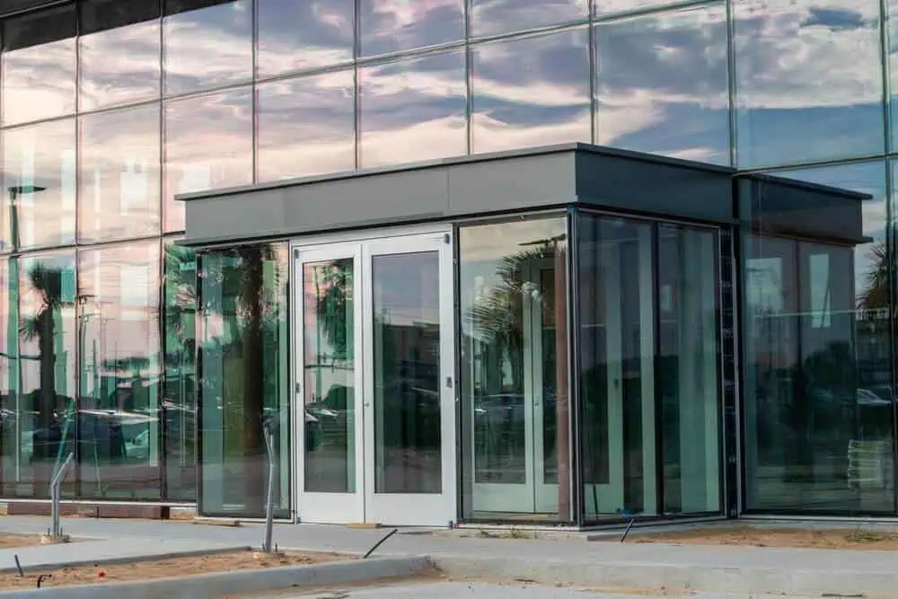 Modern glass entrance with double doors, sleek awning design, reflecting cloudy sky and lush trees, ideal for exterior renovation projects.