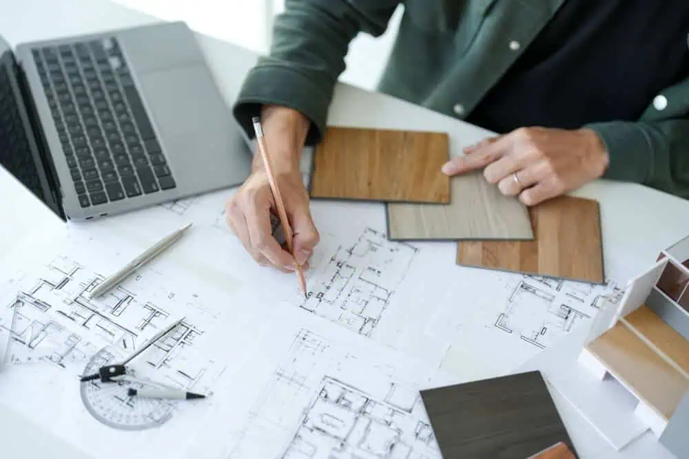 Architect working on renovation plans with laptop, wood samples, and architectural model on table.