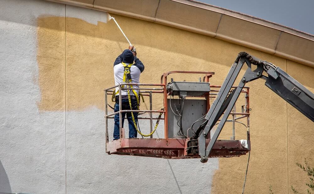 Construction worker in safety harness paints exterior wall with roller from hydraulic lift, demonstrating expert commercial painting techniques for flawless finish.