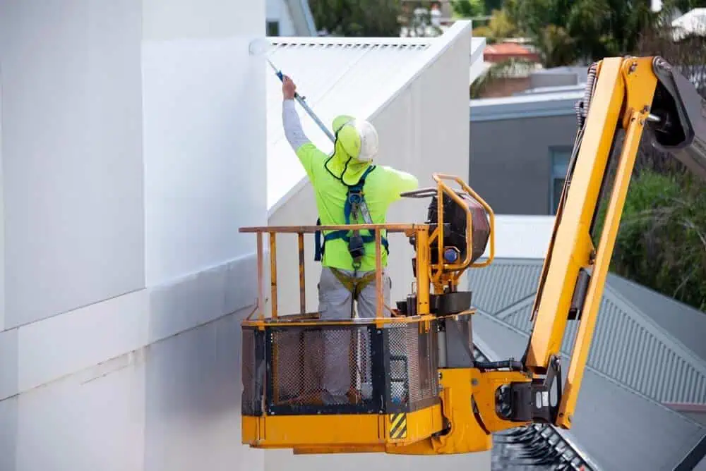 A construction worker in a safety harness painting a white wall from a yellow cherry picker, showcasing expert commercial exterior painting techniques.